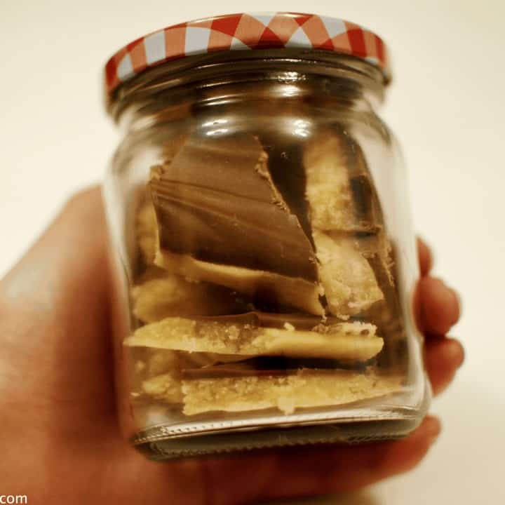 Hand holding a glass mason jar jar full of shards of caramel topped with a thin layer of milk chocolate.