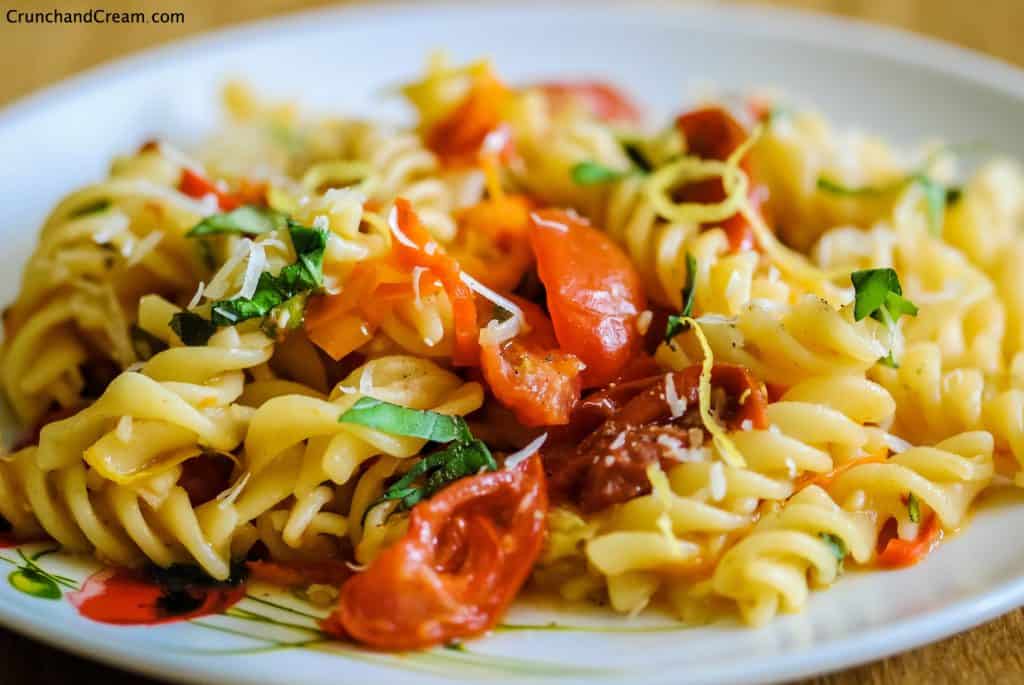 close-up eye-level of a plate with a pile of fusilli pasta with tomatoes, chopped chilli peppers, lemon zest and chopped fresh herbs