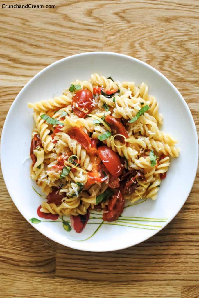 overhead image of a plate of fusilli pasta in a pile mixed with cooked tomatoes and diced chilli peppers, lemon zest and fresh herbs