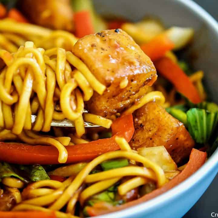 close-up of a bowl of fresh egg noodles, red bell pepper, pak choi and spring onions with crispy diced halloumi pieces in a spicy sweet chilli sauce with noodles wound around a fork