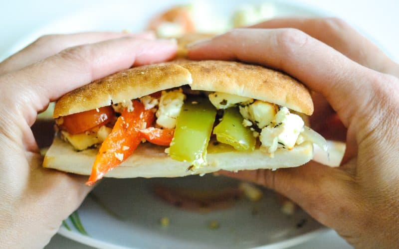 hands holding a bread roll filled with peppers, tomatoes, onions and feta