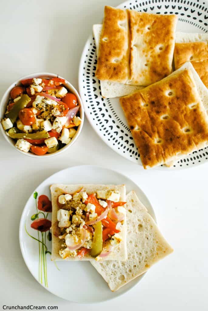 overhead of a bowl of roasted feta salad with peppers, onions and tomatoes with a plate of bread rolls. One roll is open and topped with the feta salad