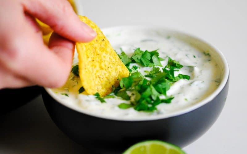 someone dunking a tortilla chip into a bowl of coriander lime dipping sauce