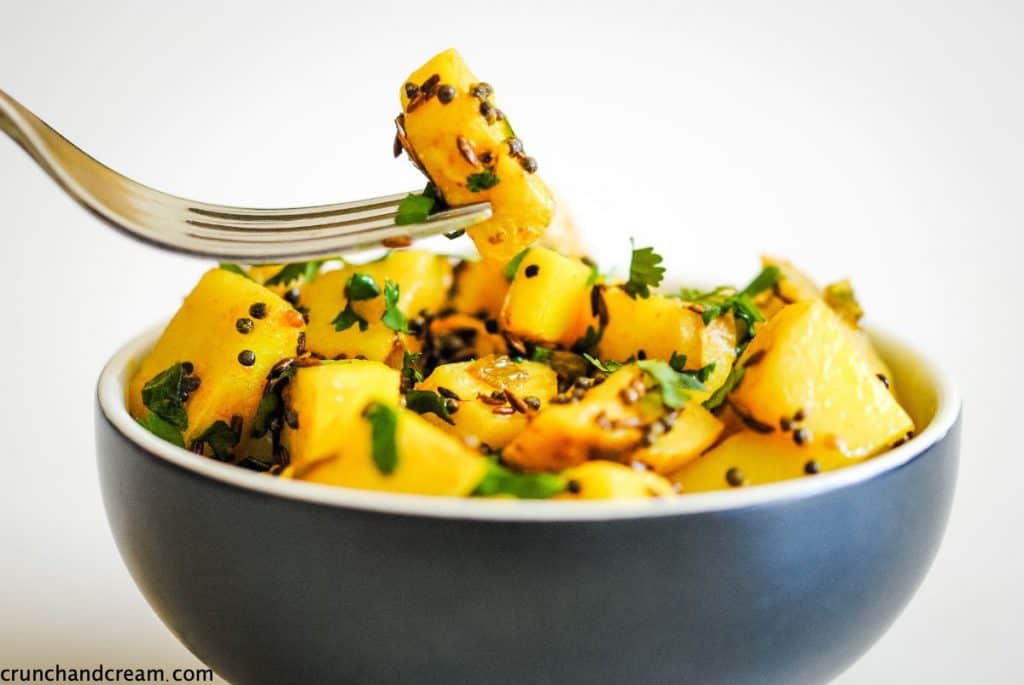 A bowl of yellow potatoes with fresh coriander and spices. A fork is holding one piece of potato.