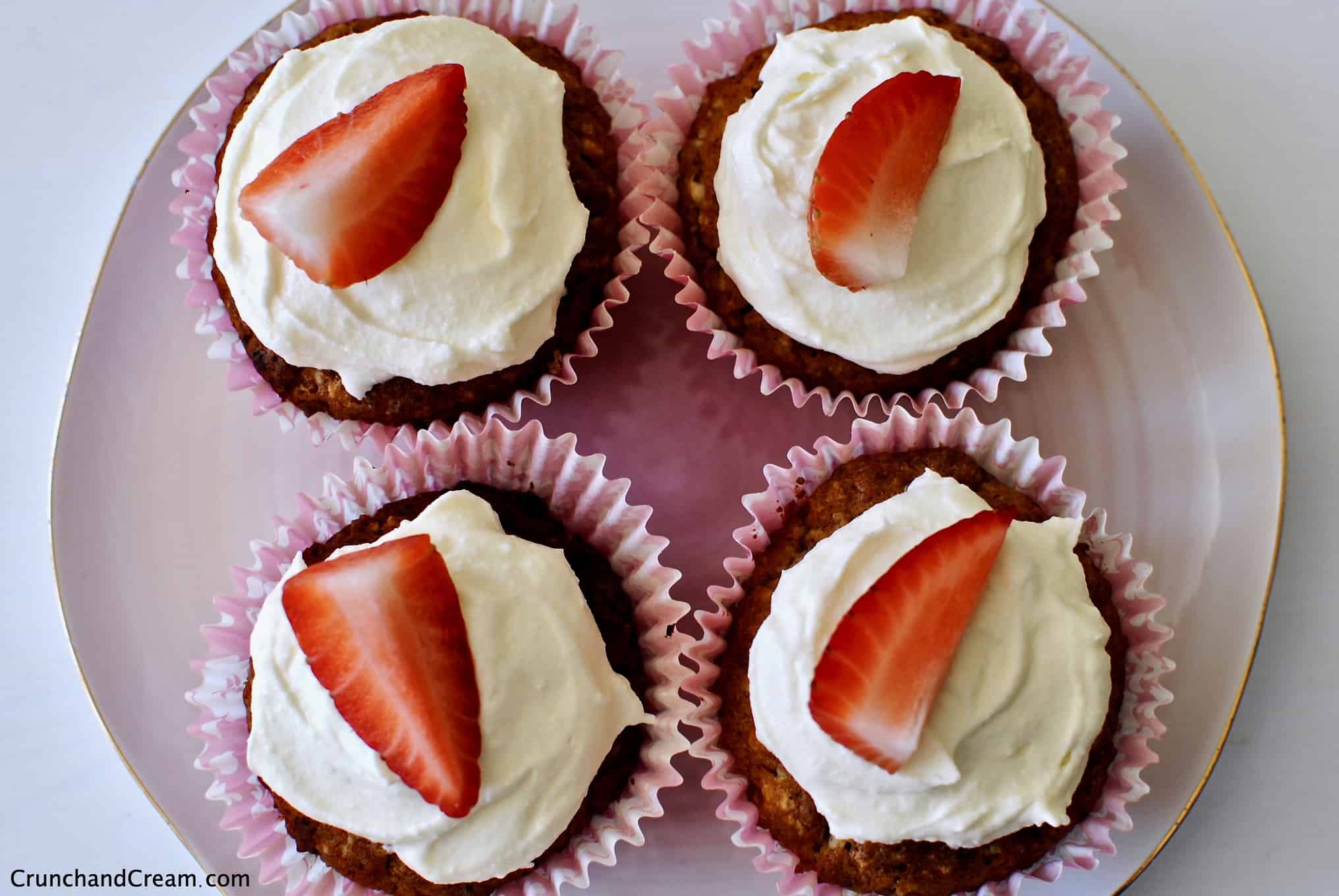 Overhead photo of 4 strawberry cupcakes topped with white chocolate cream cheese frosting and strawberry slices on a pale pink plate.